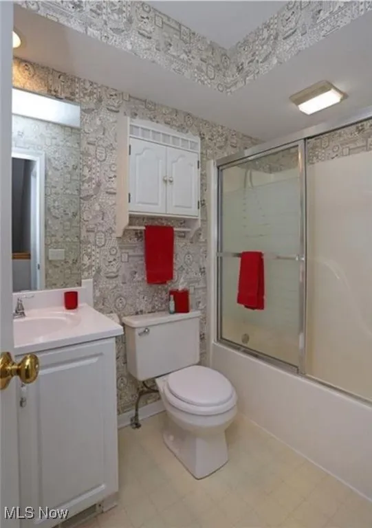 Bathroom with wallpapered walls, toilet, shower / bath combination with glass door, tile patterned flooring, and vanity