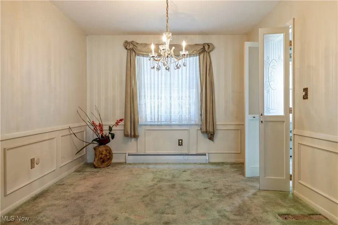 Unfurnished dining area with a baseboard heating unit, wainscoting, an inviting chandelier, a decorative wall, and carpet floors