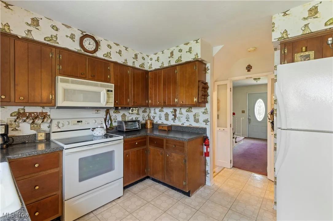 Kitchen with brown cabinetry, white appliances, dark countertops, and wallpapered walls