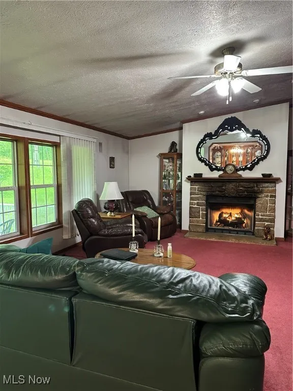 Living room with carpet, a textured ceiling, crown molding, ceiling fan, and a stone fireplace