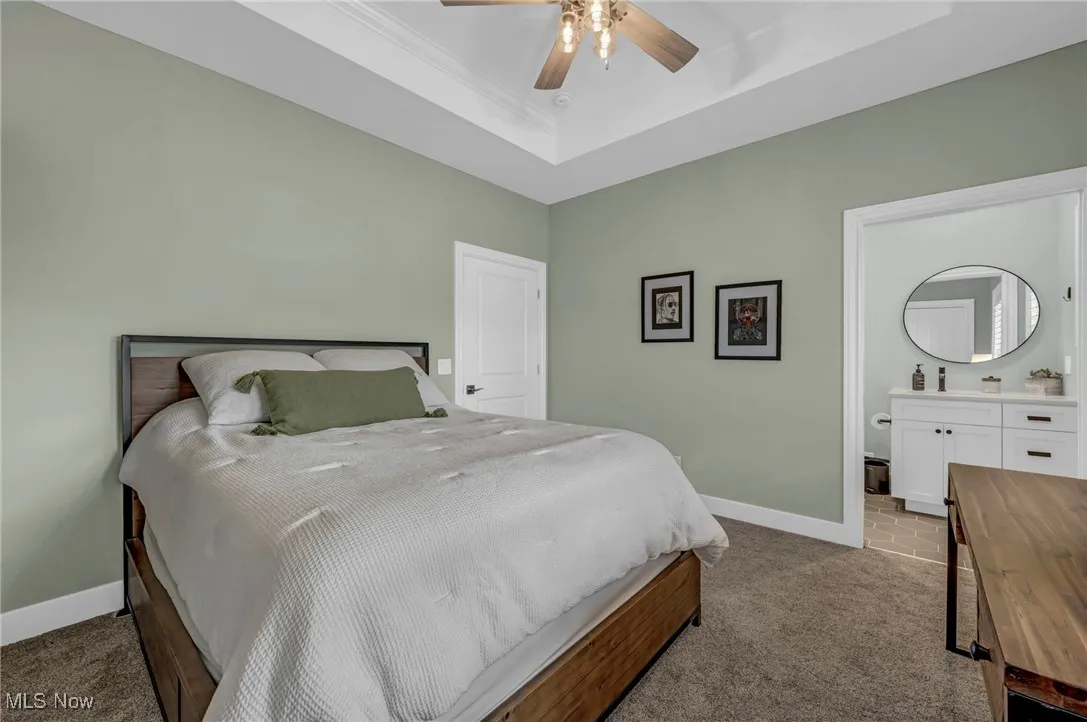 Carpeted bedroom featuring a tray ceiling, baseboards, crown molding, ensuite bath, and a ceiling fan