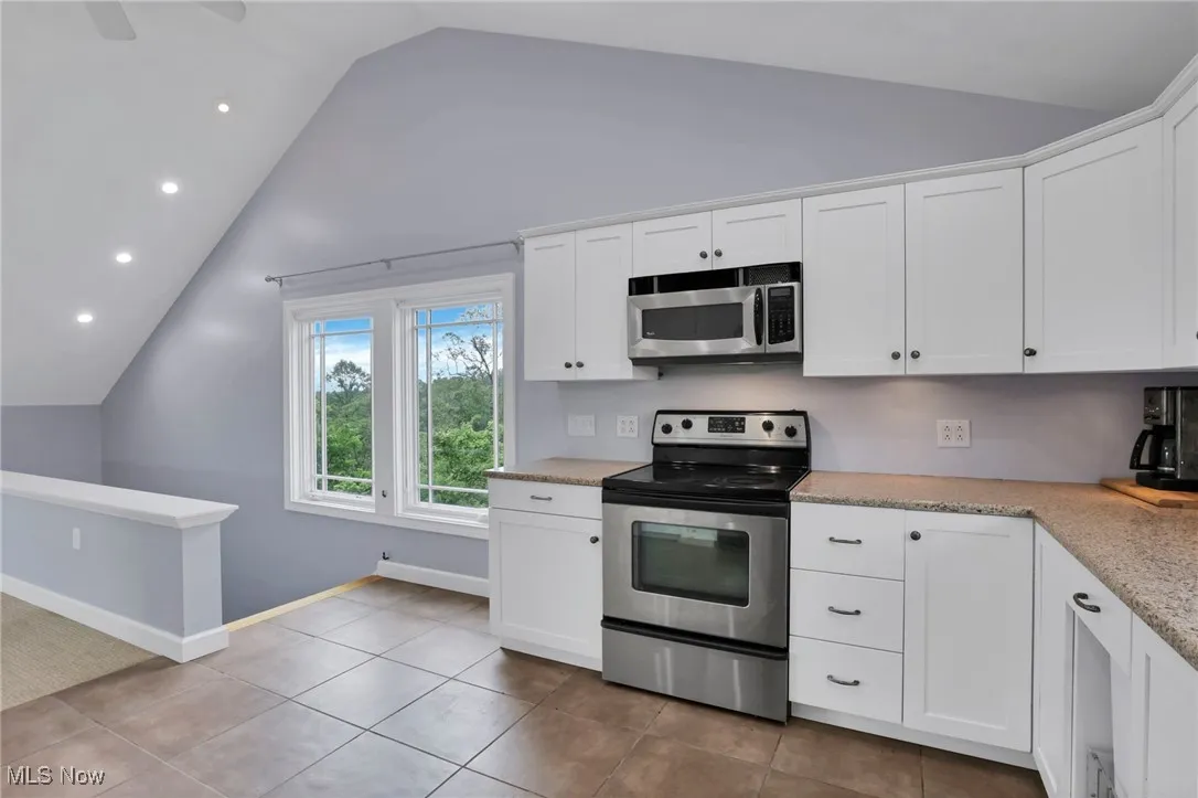 Kitchen with stainless steel appliances, vaulted ceiling, white cabinetry, light stone countertops, and light tile patterned flooring