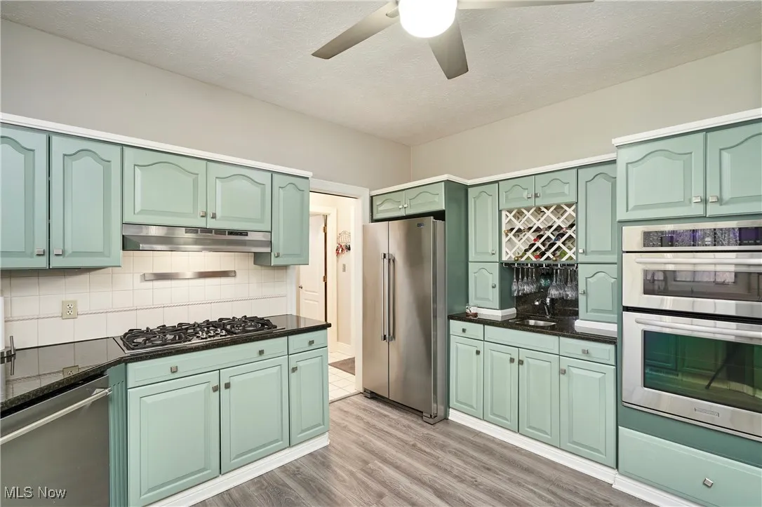 Kitchen with stainless steel appliances, under cabinet range hood, green cabinets, ceiling fan, and decorative backsplash