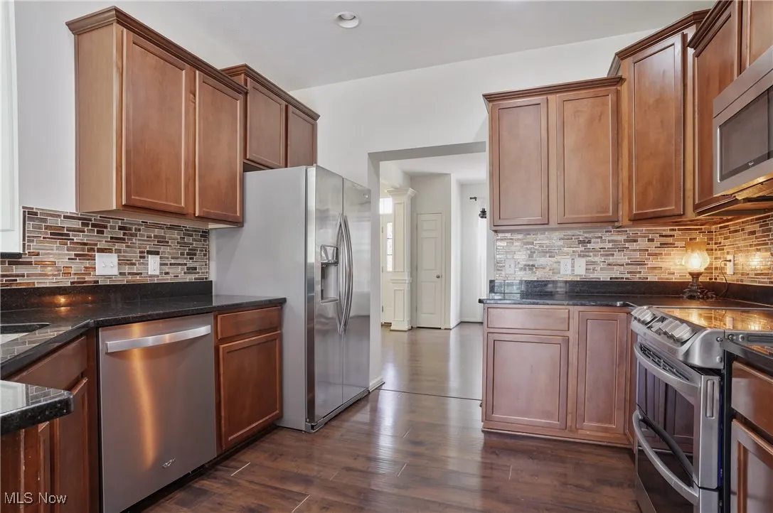 Kitchen featuring stainless steel appliances, brown cabinets, dark stone counters, dark wood finished floors, and decorative backsplash