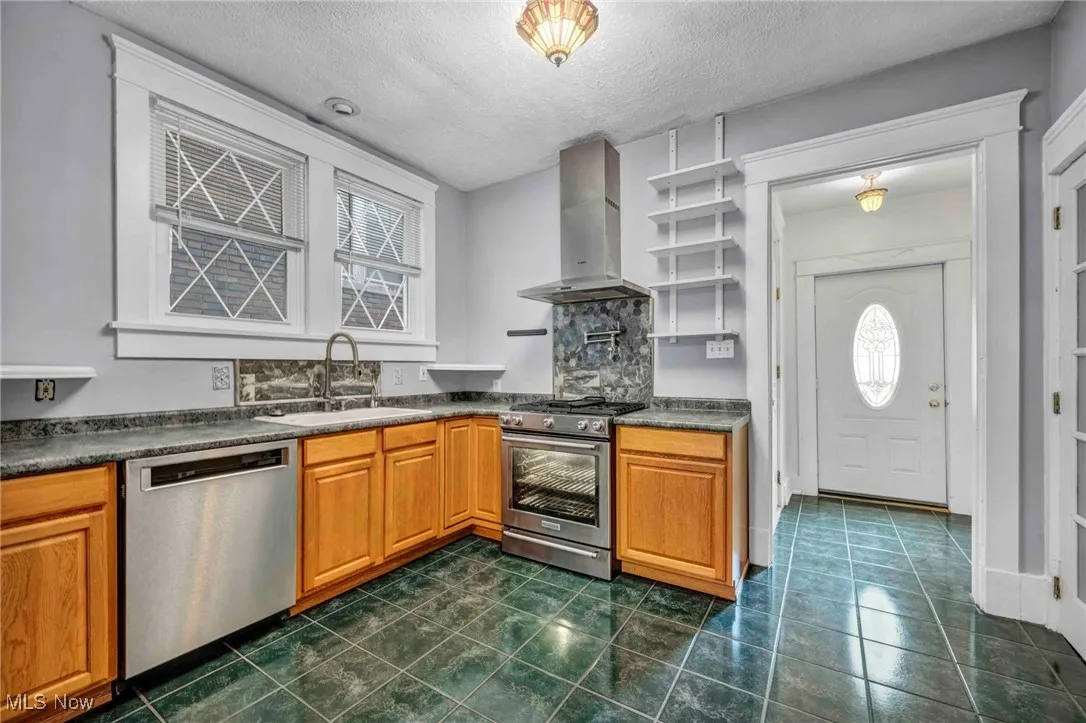 Kitchen featuring stainless steel appliances, wall chimney range hood, a sink, dark countertops, and a textured ceiling