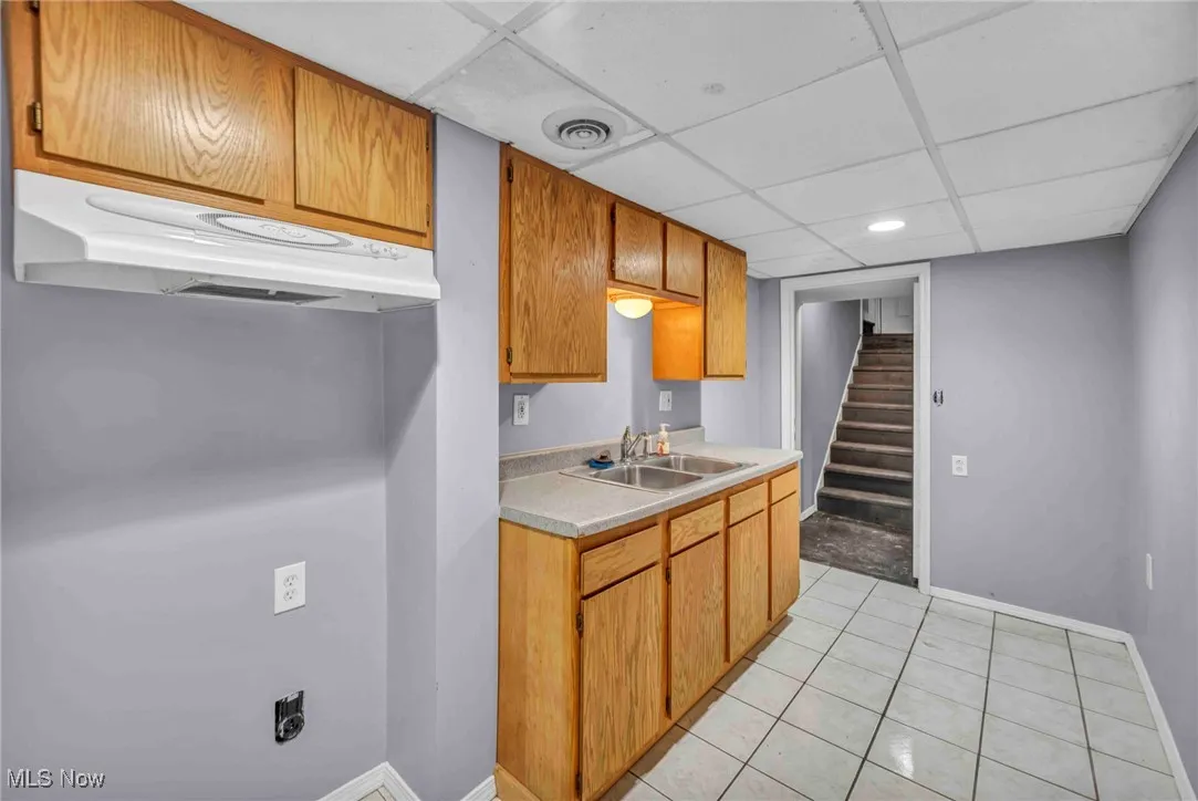 Basement Kitchen with a sink, a drop ceiling, under cabinet range hood, light countertops, and light tile patterned floors
