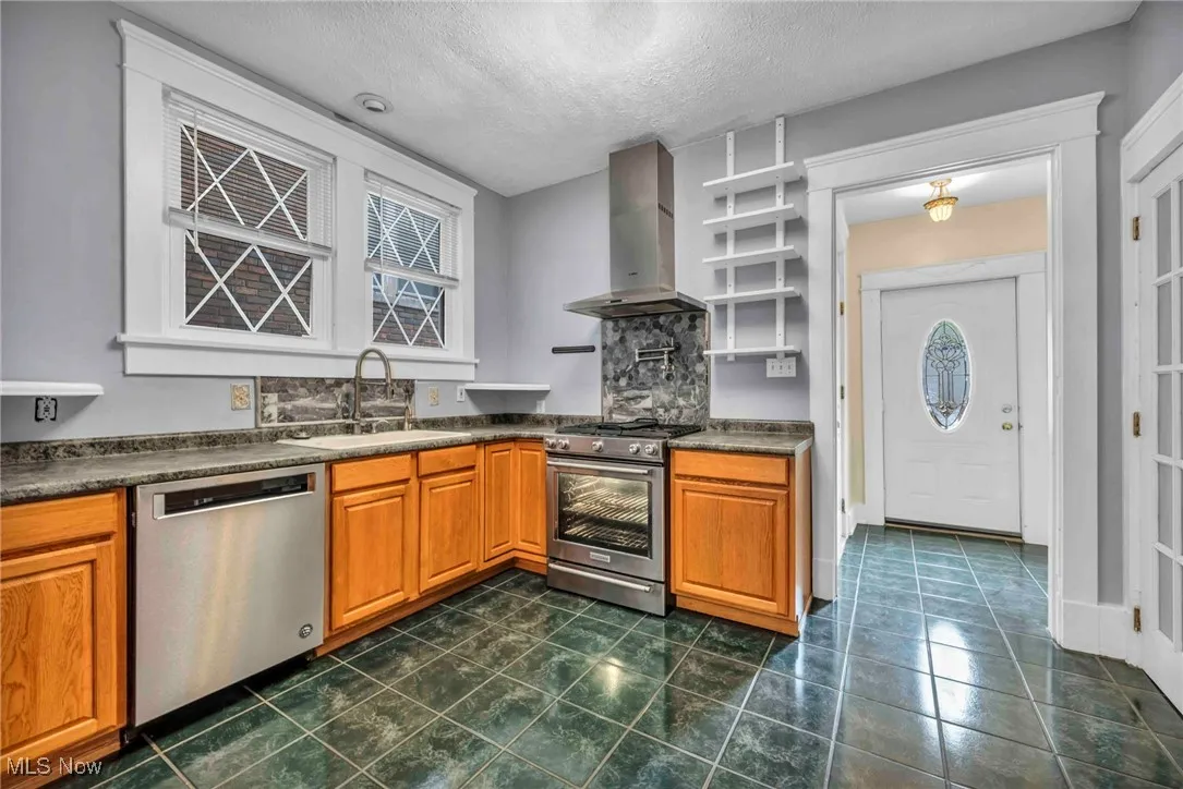 Kitchen featuring stainless steel appliances, wall chimney exhaust hood, a sink, dark countertops, and brown cabinetry