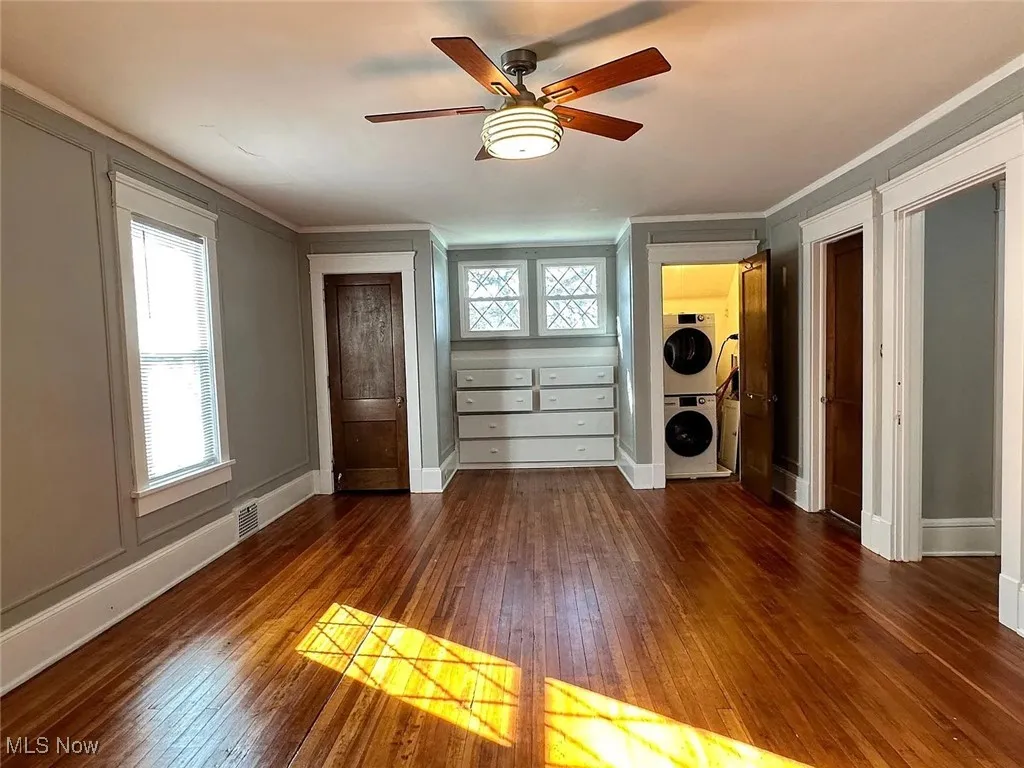 Empty room with stacked washer / drying machine, dark wood-style flooring, ornamental molding, baseboards, and ceiling fan