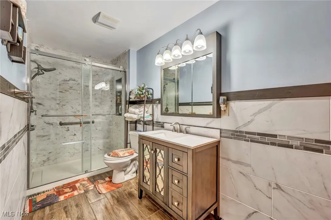 Main Floor Full bath featuring tile walls, a stall shower, toilet, vanity, and wood finished floors