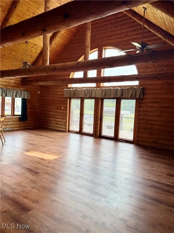 Unfurnished living room featuring a ceiling fan, a wood ceiling with exposed beams, high vaulted ceiling, wood finished floors, and rustic walls