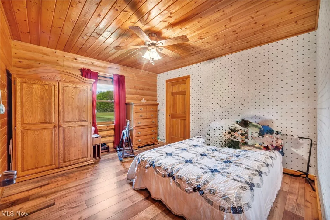 Main floorBedroom featuring wooden ceiling, light wood finished floors, wallpapered walls, ceiling fan, and log walls