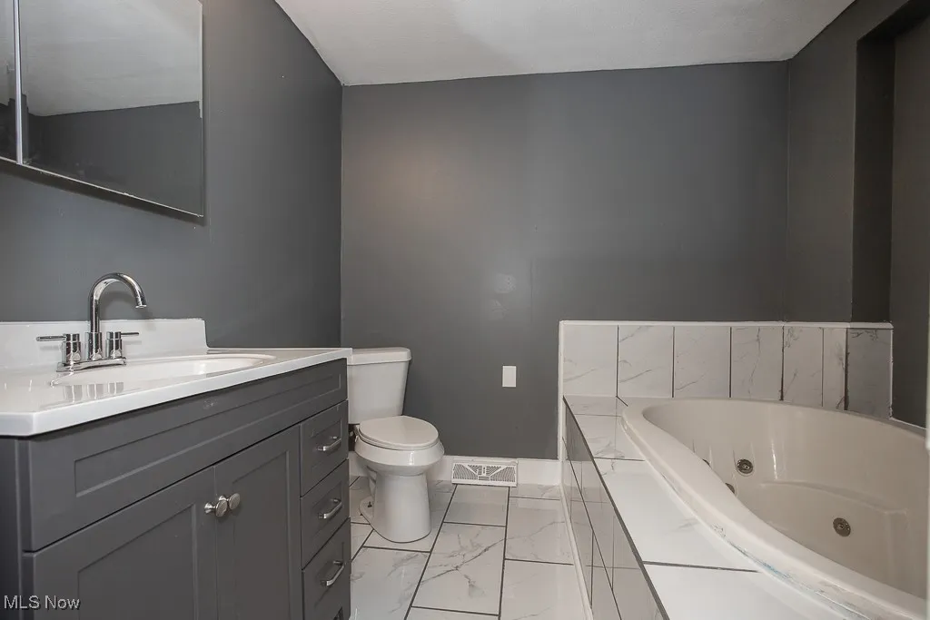 Ensuite bathroom with marble look tiles, toilet, a tub with jets, vanity, and baseboards