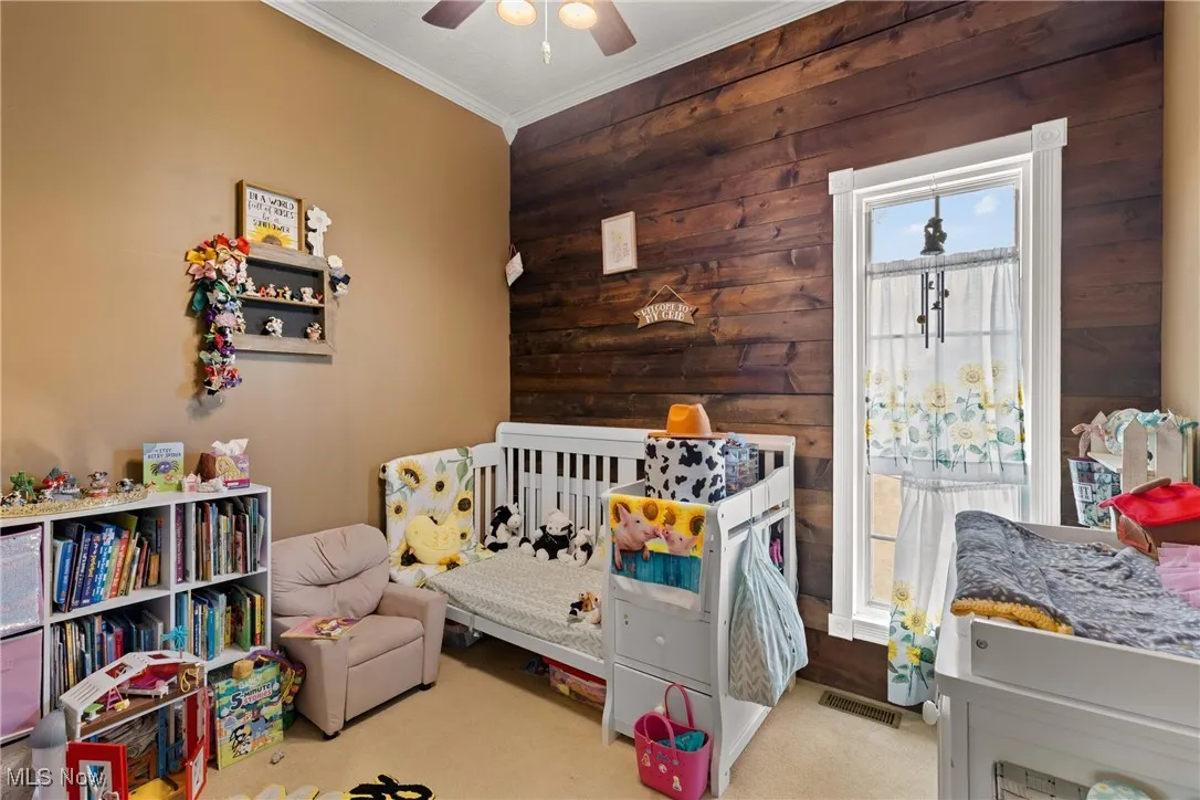 Carpeted bedroom featuring wooden walls, crown molding, a crib, and a ceiling fan