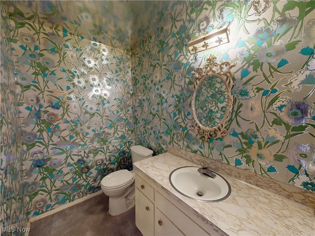 Half bathroom featuring wallpapered walls, baseboards, toilet, and vanity