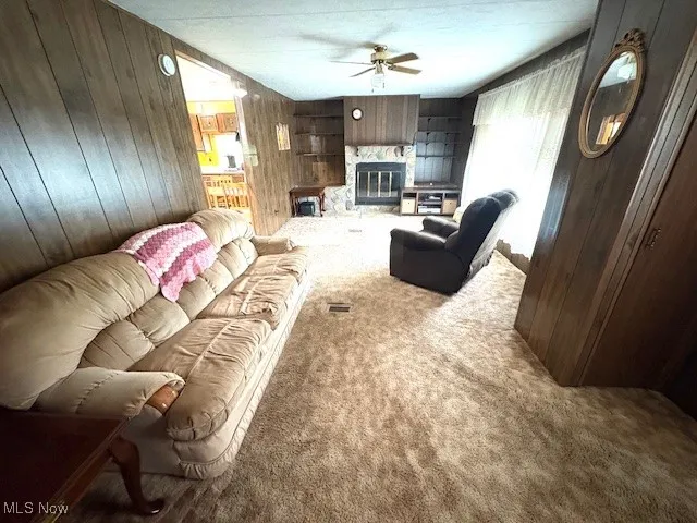 Carpeted living room with a ceiling fan, wood walls, and a stone fireplace