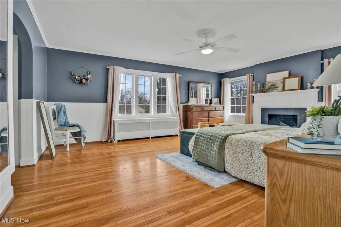 Bedroom with wainscoting, a brick fireplace, light wood-style flooring, arched walkways, and a ceiling fan