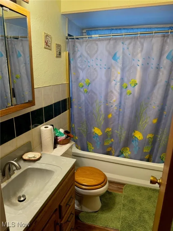 Full bathroom with tile walls, vanity, shower / bath combo with shower curtain, a textured wall, and wainscoting