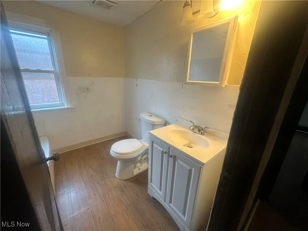 Half bath with wood finished floors, toilet, vanity, tile walls, and wainscoting