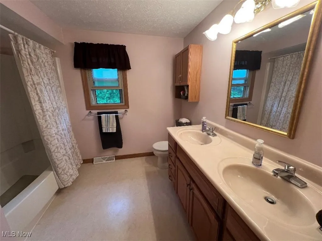 Full bath featuring toilet, double vanity, a textured ceiling, baseboards, and shower / bath combo