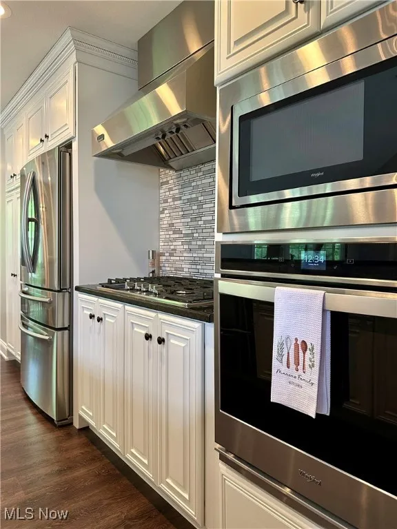 Kitchen featuring appliances with stainless steel finishes, wall chimney range hood, dark countertops, backsplash, and white cabinets
