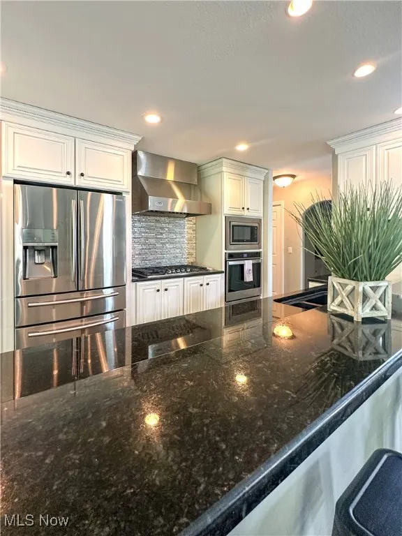 Kitchen with stainless steel appliances, wall chimney exhaust hood, backsplash, dark stone countertops, and white cabinets