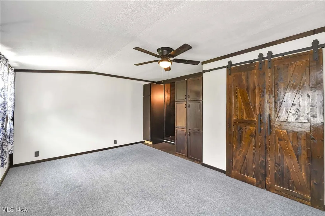 Unfurnished bedroom featuring carpet, a barn door, baseboards, crown molding, and ceiling fan