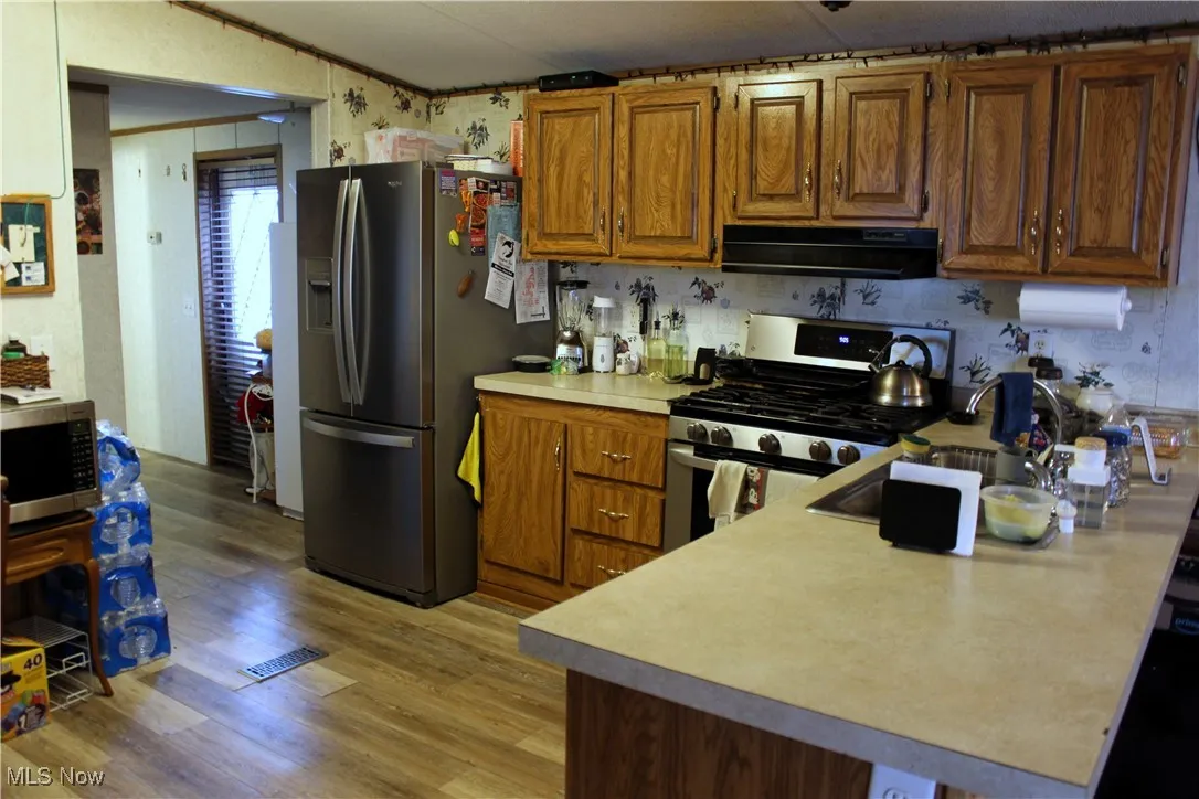 Kitchen with appliances with stainless steel finishes, ventilation hood, brown cabinets, light wood-style floors, and light countertops