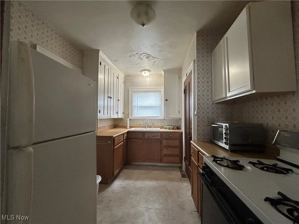 Kitchen with white appliances, wallpapered walls, a sink, light countertops, and brown cabinetry
