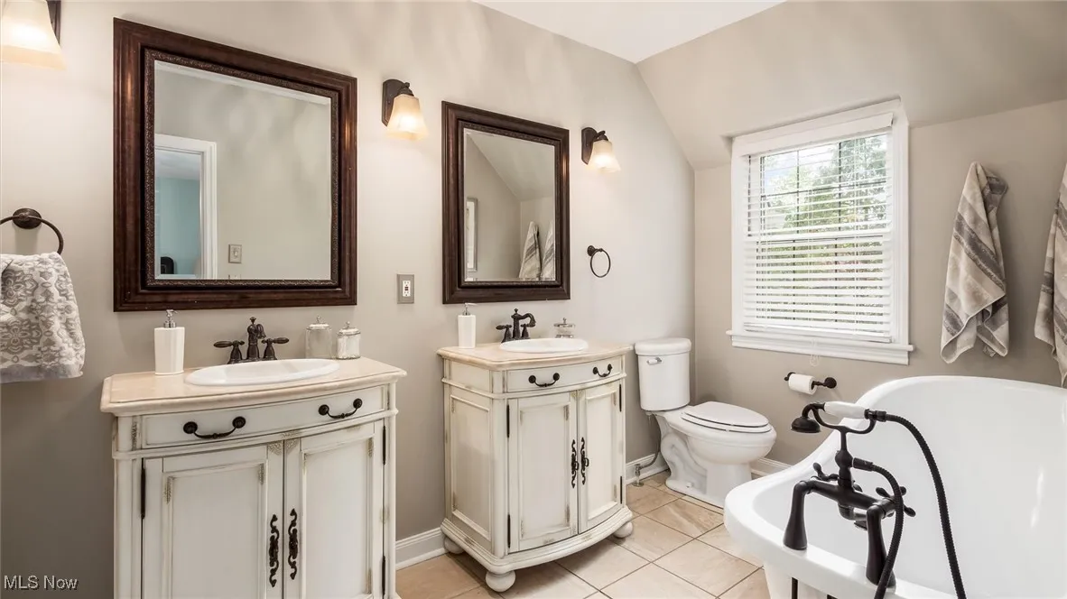 Bathroom with a soaking tub, baseboards, two vanities, tile patterned flooring, and toilet