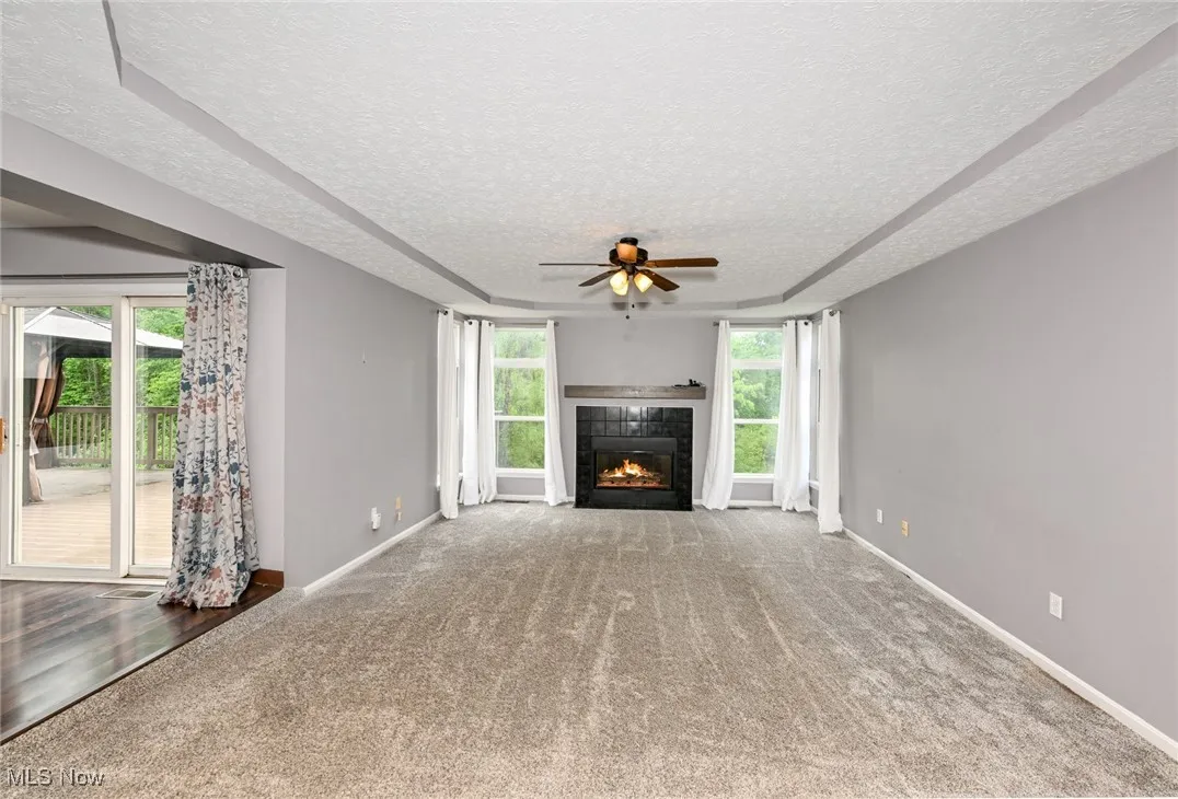 Unfurnished living room with a textured ceiling, carpet, a ceiling fan, a tiled fireplace, and baseboards adjacent to kitchen
