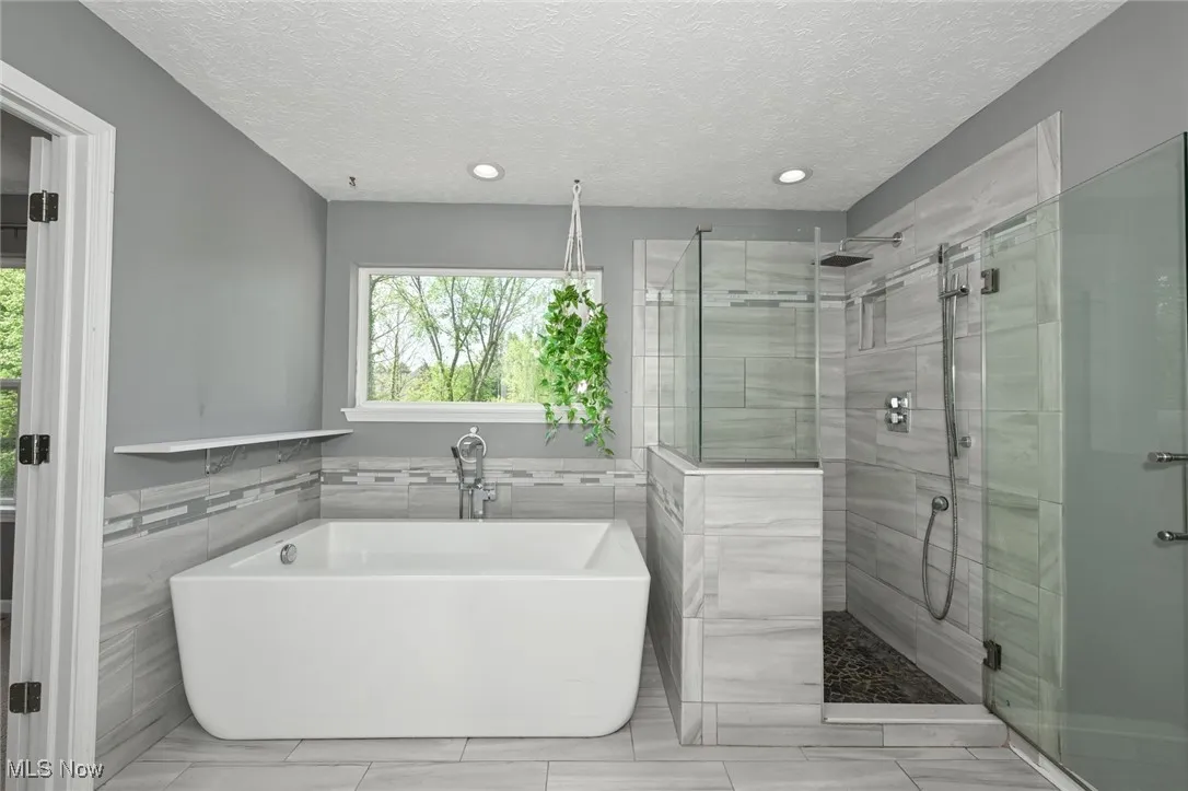 Full updated bath featuring tile walls, a stall shower, a freestanding tub, a textured ceiling, and recessed lighting