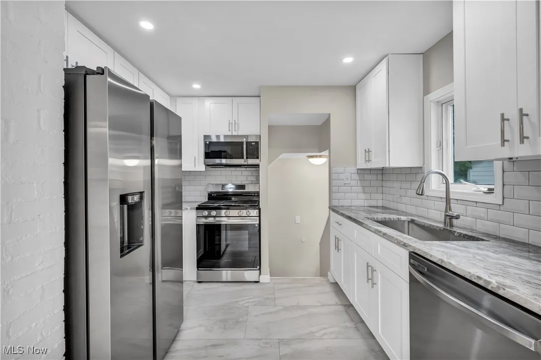 Kitchen featuring appliances with stainless steel finishes, a sink, light marble finish floors, light stone countertops, and white cabinets