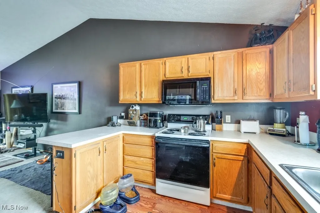 Kitchen with electric range, black microwave, lofted ceiling, light countertops, and a sink