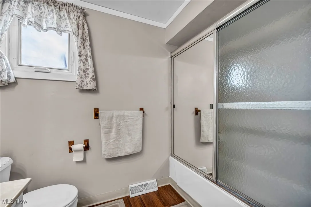 Full bath with toilet, crown molding, bath / shower combo with glass door, wood finished floors, and baseboards
