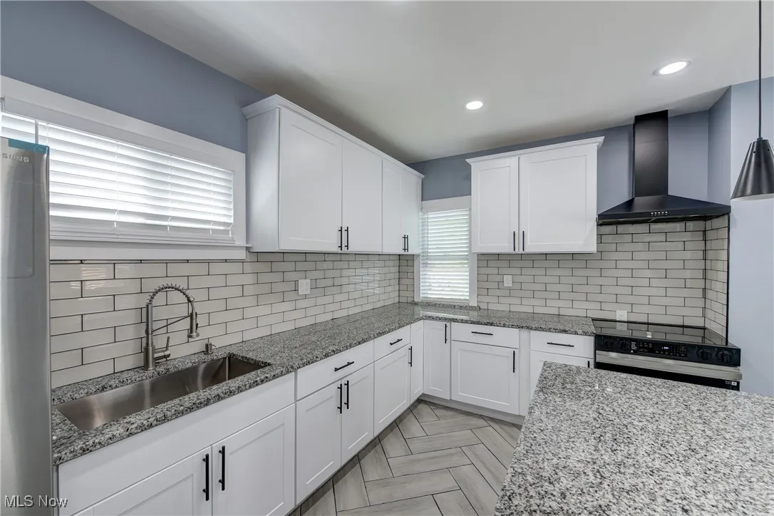Kitchen with wall chimney range hood, a sink, white cabinets, light stone countertops, and recessed lighting