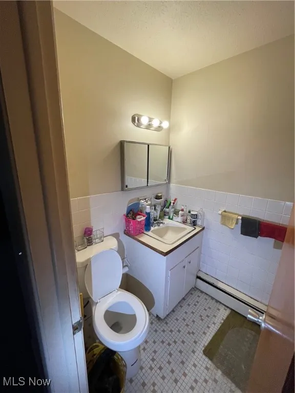 Bathroom with tile walls, a baseboard radiator, toilet, a wainscoted wall, and vanity