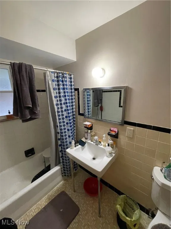 Full bathroom with tile walls, toilet, shower / bathtub combination with curtain, and wainscoting