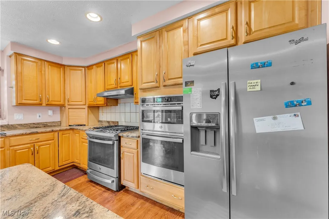 Kitchen with stainless steel appliances, under cabinet range hood, light wood-style flooring, light stone countertops, and decorative backsplash