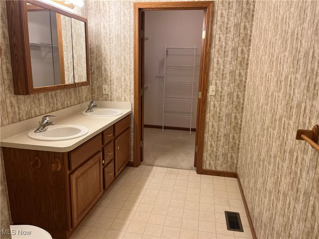 Full bath featuring wallpapered walls, baseboards, a walk in closet, double vanity, and tile patterned floors