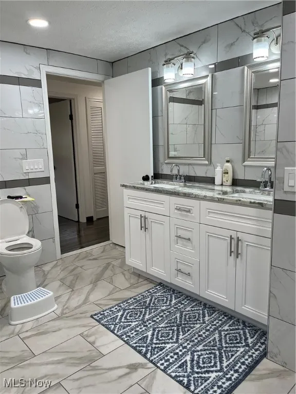 Full bath with marble look tile flooring, tile walls, double vanity, toilet, and a textured ceiling