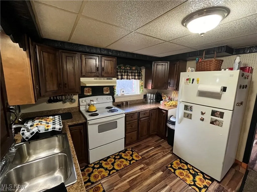 Kitchen with white appliances, under cabinet range hood, a sink, dark wood finished floors, and a paneled ceiling