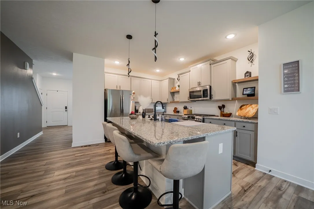 Kitchen with stainless steel appliances, open shelves, wood finished floors, a center island with sink, and light stone countertops