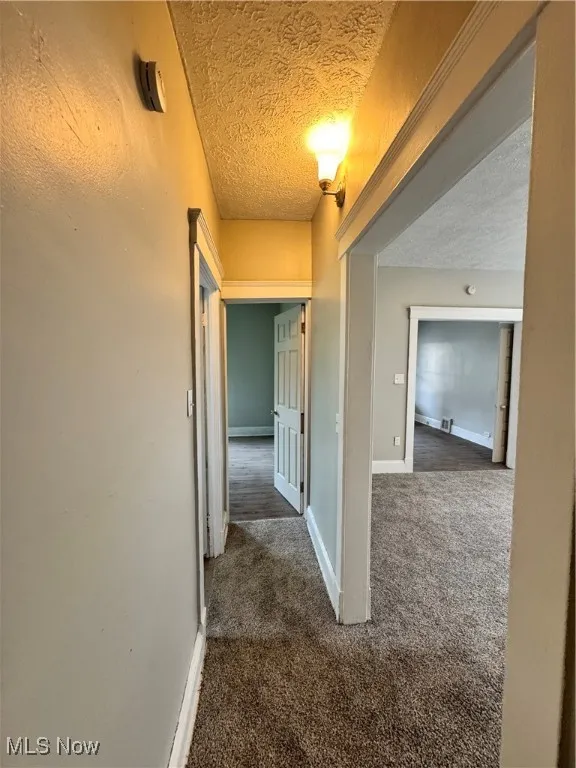 Hallway featuring a textured ceiling, dark colored carpet, and baseboards