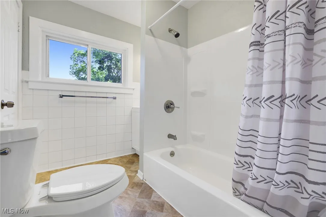 Bathroom  toilet, shower / bathtub combination with curtain, stone finish flooring, and wainscoting