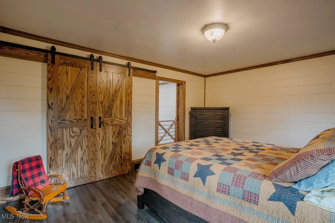 Bedroom with dark wood-type flooring, wood walls, a barn door, and ornamental molding