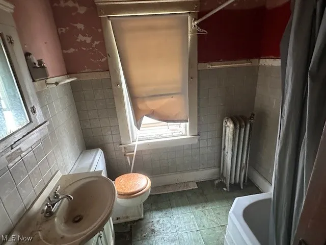 Bathroom with radiator heating unit, tile walls, toilet, a sink, and a wainscoted wall