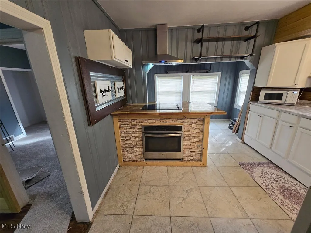 Kitchen with stainless steel oven, wall chimney range hood, white microwave, white cabinetry, and wooden walls