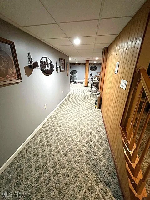 Hall with a drop ceiling, carpet flooring, baseboards, and wood walls