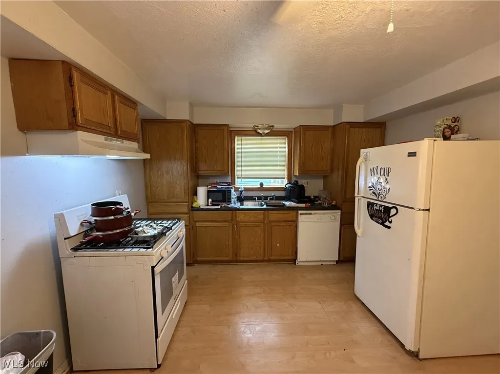 Kitchen with a textured ceiling, brown cabinets, a sink, under cabinet range hood, and white appliances