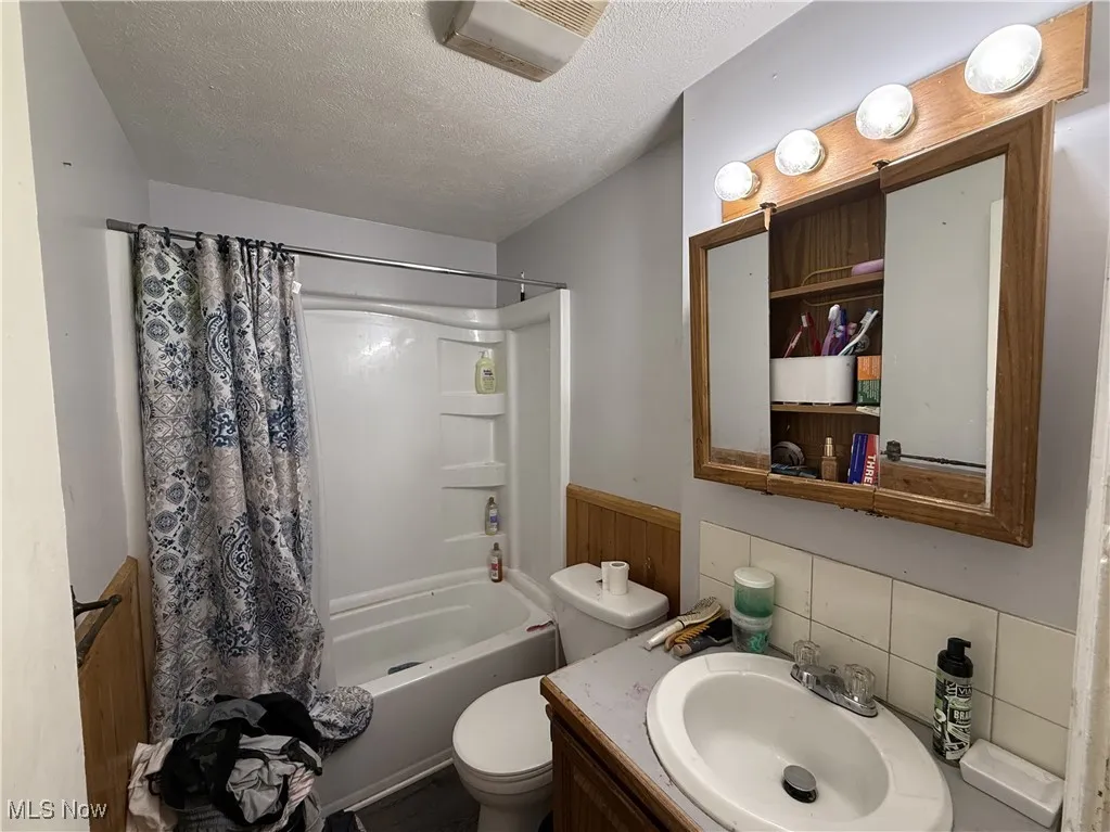 Full bathroom with shower / bathtub combination with curtain, a wainscoted wall, toilet, vanity, and a textured ceiling